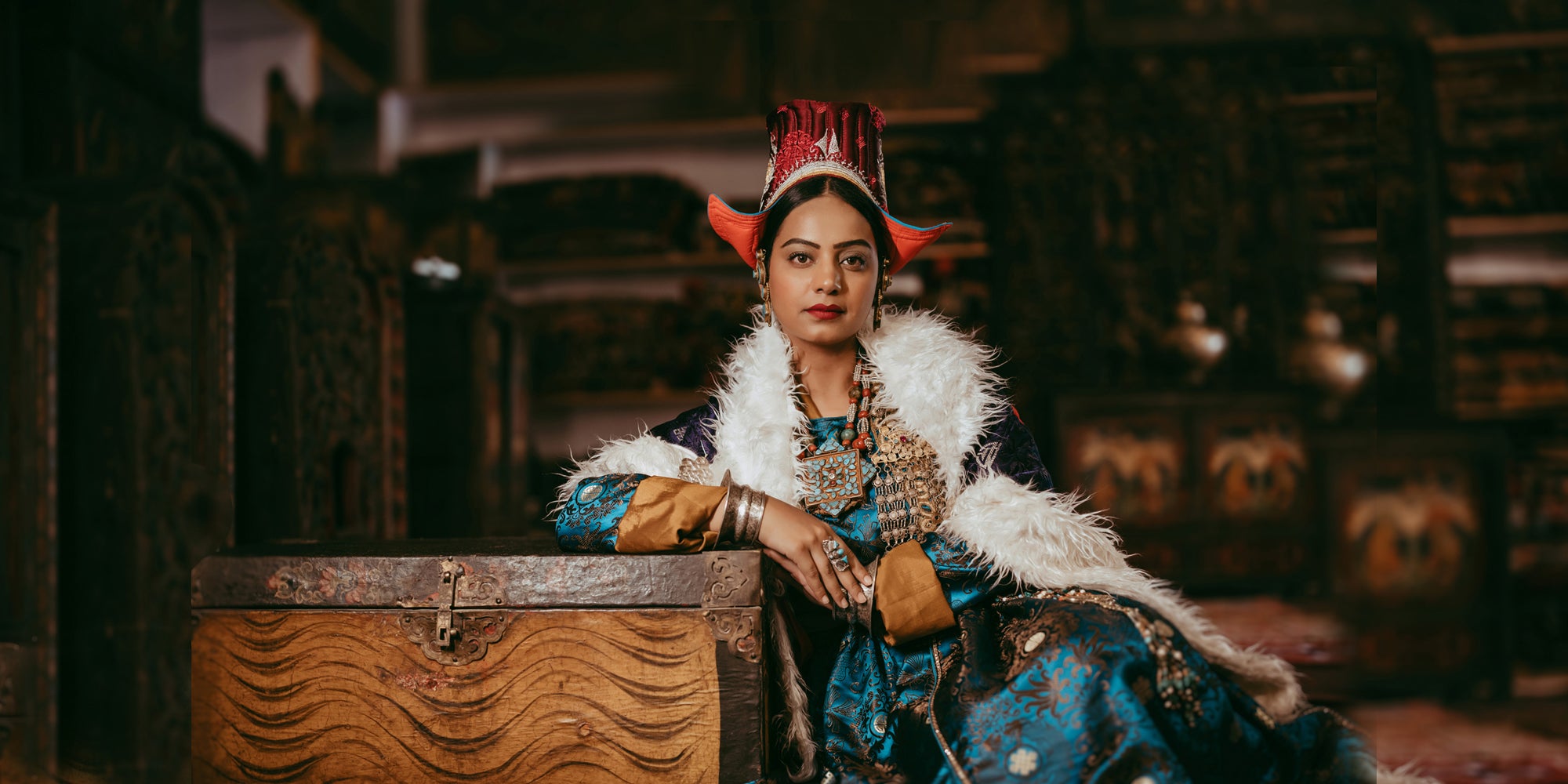 Person in traditional attire with decorative headdress and fur, sitting next to a wooden chest.